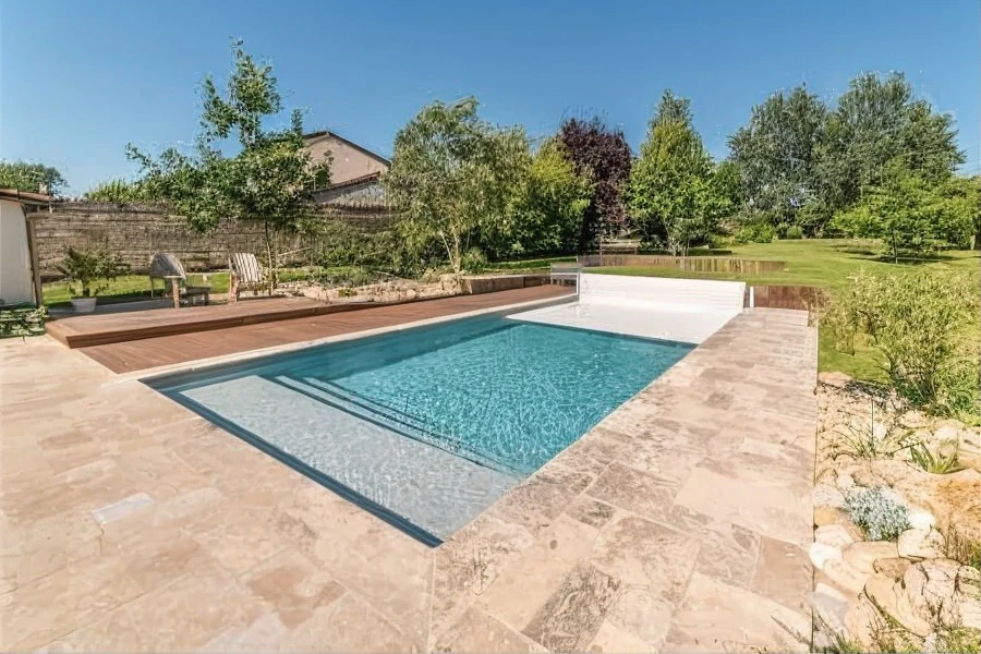 Piscine extérieure résidentielle avec cascade en pierre naturelle, terrasse en pavés, mobilier de jardin noir, aménagement paysager luxueux avec arbres ornementaux rouges et verts, éclairage décoratif suspendu