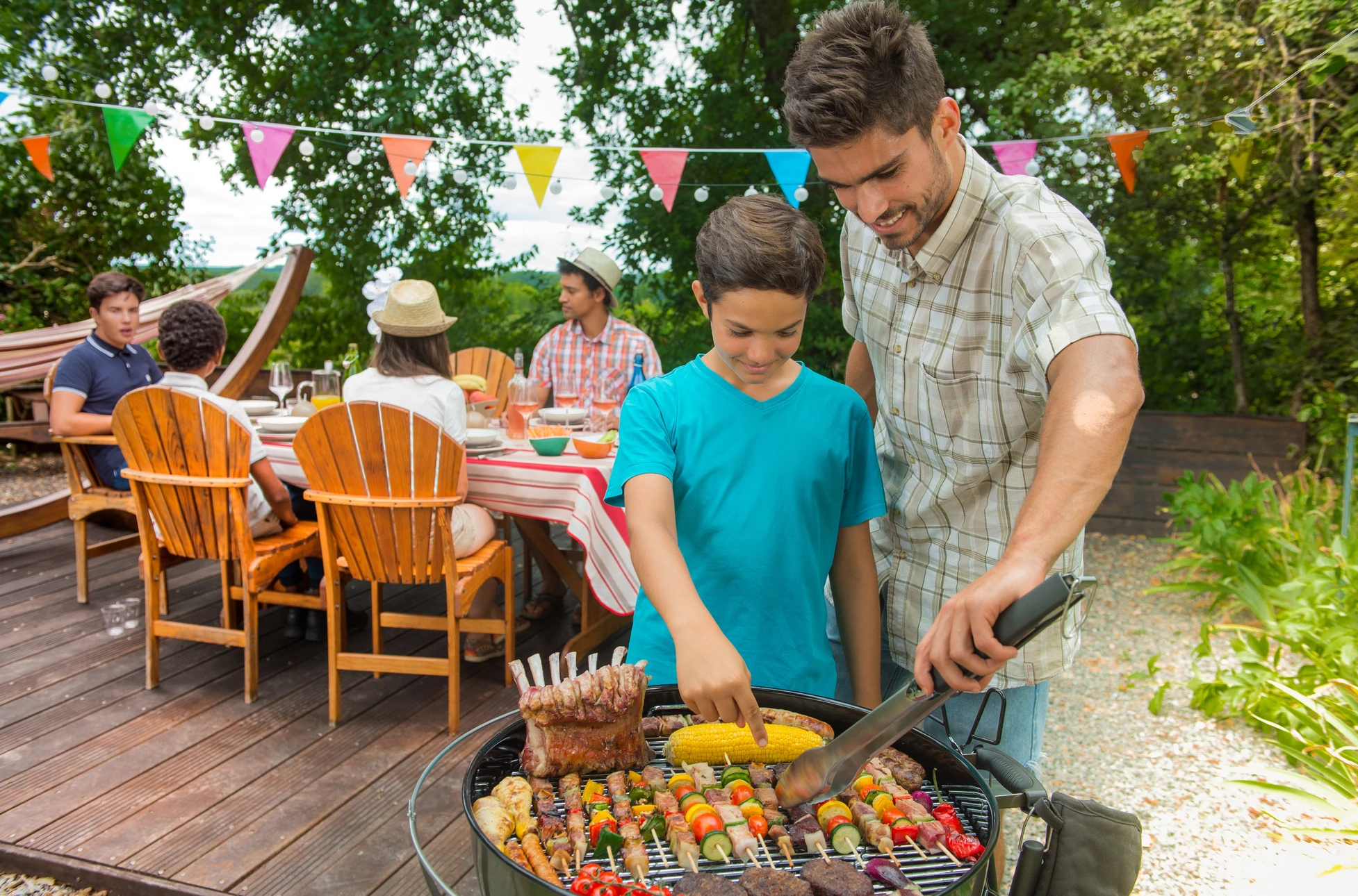 Scène extérieure de barbecue familial sur terrasse en bois avec père et fils cuisinant ensemble