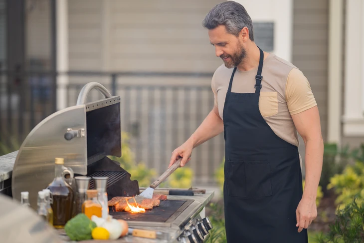 Homme d'âge moyen avec tablier noir grillant de la viande sur barbecue à gaz en extérieur, spatule à la main, souriant