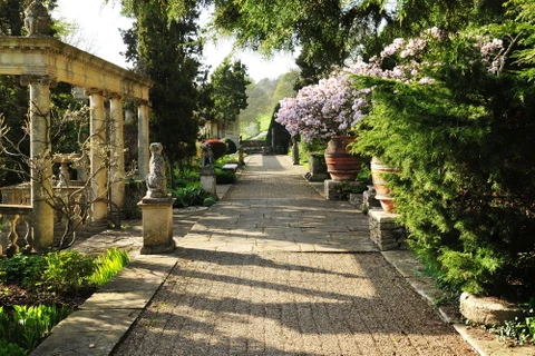 Jardin paysager avec allée pavée bordée de colonnes classiques en pierre calcaire, statues ornementales et grands pots en terre cuite garnis de végétaux fleuris