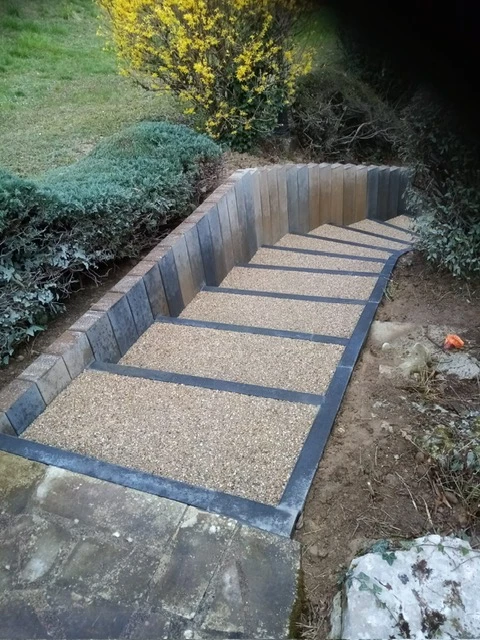Jardin paysager avec allée pavée bordée de colonnes classiques en pierre calcaire, statues ornementales et grands pots en terre cuite garnis de végétaux fleuris